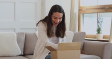 Excited Young Woman Customer Opening Parcel Box At Home. Amazed Happy Girl Shopper Unboxing Fashion Purchase Sitting On Couch. Satisfied Female Consumer Unpacking Postal Shipping Delivery Concept
