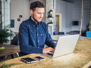 happy smiling remote online working man typing and looking into laptop screen with mobile phone and notebook in casual outfit standing up in front of a work desk in an coworking office