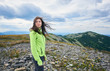 © anatoliy_gleb - Young female with hair flying from the wind is looking into distance against backdrop of mountains valley. Among the stones yellow mountain flowers break through under cloudy sky.Girl in bright jacket