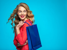 Cheerful  Young Woman Holds Bags With A Purchases. Joyful Trendy Girl With A Colorful Shopping Bags. Model In A Red Shirt Over Blue Background Holding Bags With Gifts. Happy Caucasian Woman.