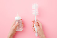 Young Mother Hands Holding Baby Milk Bottle And White Brush On Pastel Pink Table Background. Preparing For Washing. Closeup. Point Of View Shot. Top Down View.