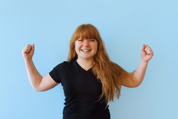 Pretty Caucasian red-haired woman keeps herself fit and healthy, raises her hands and shows muscles, takes pride in her achievements in the gym, smiles broadly, dressed in black casual clothes