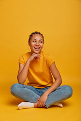 Wall Mural - Vertical photo of good looking cheerful dark skinned lady keeps hand under chin, looks aside with broad smile, sits crossed legs on floor against yellow background, enjoys spare time with friends