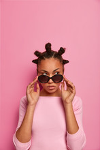 Beauty Feminine Fashion Model Wears Stylish Sunglasses, Looks Thoughtfully And With Suspicion, Has Bun Hairstyle, Isolated On Pastel Rosy Wall, Empty Space Above For Your Promotion. Studio Shot