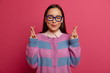 Hopeful optimistic woman crosses fingers, anticipates results, eager to fullfill dream, closes eyes, wears big optical spectacles and jumper, poses against pink background, pleads and makes wish