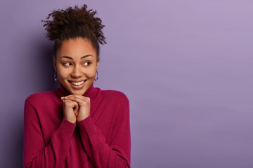 Wall Mural - Dreamy happy ethnic woman has interesting idea, keeps hands under chin and looks aside, thinks about something good, dreams about vacation with boyfriend, stands over purple wall, blank space