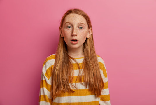 Shocked ginger freckled girl stands with opened mouth, stands speechless and reacts on stunning news, dressed in striped casual sweater, poses against pink background. Oh my God what happened