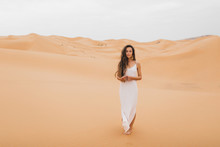 Pretty Portrait Of Young Beautiful Woman In Sand Dunes Of Moroccan Sahara Desert. Brunette With Long Hair, Eastern Appearance. Bride In White Silk Dress.