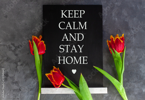 Words Keep Calm And Stay Home on chalkboard on grey concrete background with fresh tulips. Virus quarantine banner against COVID-19 coronavirus. Blooming flowers flat lay card. Text sign black board