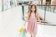 © etonastenka - Portrait of a little happy girl in the mall. A smiling laughing girl in a pink dress with a cute rim with ears and with multi-colored bags in her hands is walking around the mall, looking at the