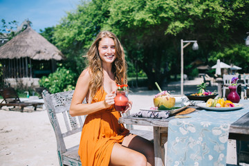 Cheerful adult woman having nutrition while sitting at table on sea sand beach in cafe