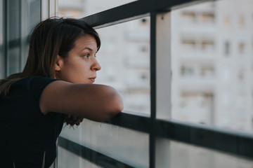 Young sad woman looking outside through balcony of an apartment building