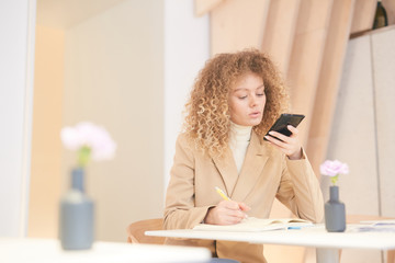 Wall Mural - Warm-toned portrait of curly-haired young woman dictating message via smartphone while working or studying sitting at table in cafe, copy space