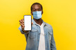 © khosrork - Online service, technology. Portrait of satisfied glad young man with medical mask standing holding out cellphone and smiling broadly at camera. indoor studio shot isolated on yellow background
