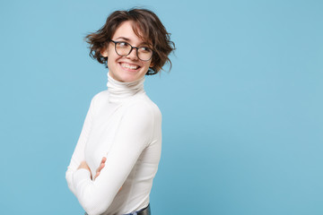 Side view of smiling young woman girl in casual white clothes, eyeglasses posing isolated on pastel blue background. People lifestyle concept. Mock up copy space. Holding hands crossed, looking aside.