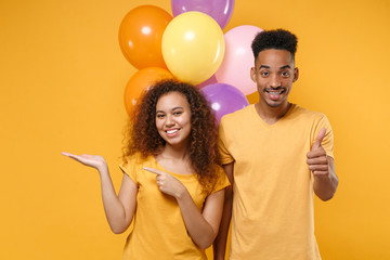 Poster - Smiling friends couple african american guy girl isolated on yellow background. Birthday holiday party concept. Celebrate hold colorful air balloons pointing index finger hand aside, showing thumb up.