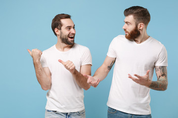 Excited two young men guys friends in white t-shirt posing isolated on pastel blue background. Sport leisure lifestyle concept. Cheer up support favorite team pointing thumbs aside, spreading hands.