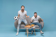 © ViDi Studio - Excited young men guys friends in white t-shirt sit on couch isolated on pastel blue background. Sport leisure concept. Cheer up support favorite team with soccer ball screaming doing winner gesture.