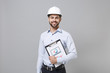 © ViDi Studio - Smiling young unshaven business man in light shirt, protective construction white helmet isolated on grey background. Achievement career wealth business concept. Hold clipboard with papers document.