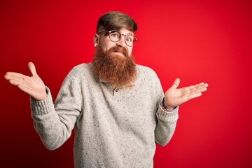 Wall Mural - Handsome Irish redhead man with beard wearing casual sweater and glasses over red background clueless and confused expression with arms and hands raised. Doubt concept.