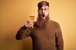 Irish redhead man with beard drinking a glass of refreshing beer over yellow background scared in shock with a surprise face, afraid and excited with fear expression