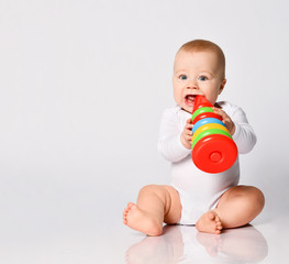 Wall Mural - Little child in bodysuit, barefoot. He chewing multi-colored stacking rings toy, sitting on floor isolated on white. Close up