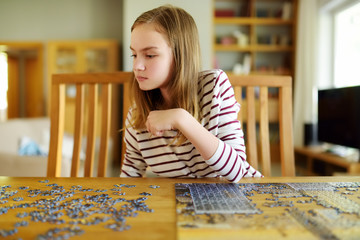 Wall Mural - Cute young girl playing puzzles at home. Child connecting jigsaw puzzle pieces in a living room table. Kid assembling a jigsaw puzzle. Fun family leisure.