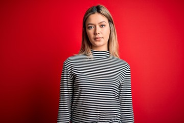 Canvas Print - Young beautiful blonde woman wearing casual striped sweater over red isolated background Relaxed with serious expression on face. Simple and natural looking at the camera.