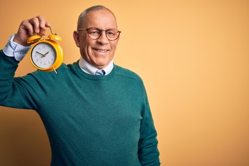 Senior handsome man holding alarm clock standing over isolated yellow background with a happy face standing and smiling with a confident smile showing teeth