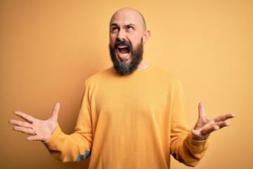 Poster - Handsome bald man with beard wearing casual sweater standing over yellow background crazy and mad shouting and yelling with aggressive expression and arms raised. Frustration concept.