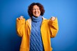 © Krakenimages.com - Middle age curly hair woman wearing rain coat standing over isolated blue background very happy and excited doing winner gesture with arms raised, smiling and screaming for success. Celebration