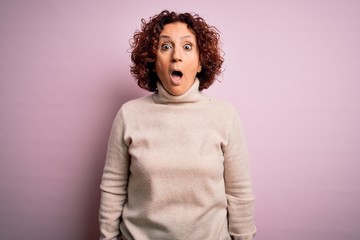 Poster - Middle age beautiful curly hair woman wearing casual turtleneck sweater over pink background afraid and shocked with surprise and amazed expression, fear and excited face.