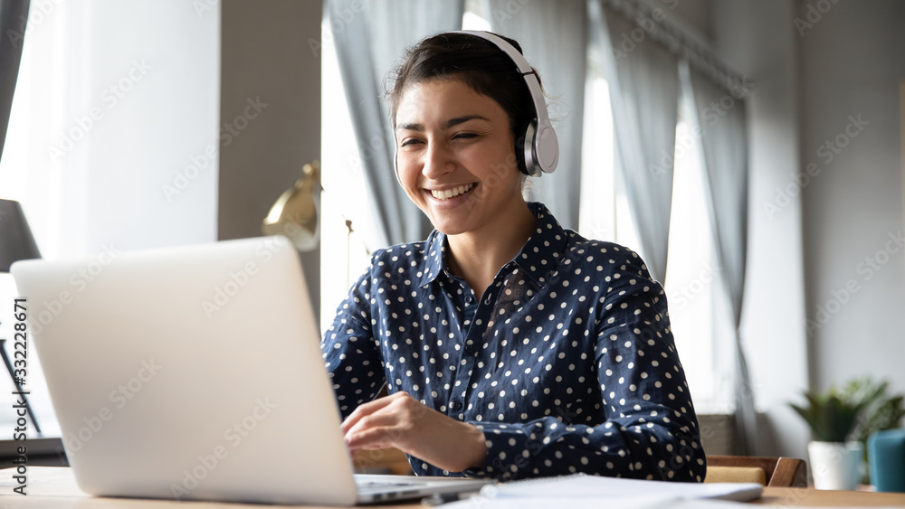 Smiling Indian girl wearing headphones using laptop, looking at screen, happy young female listening to favorite music while working online on project, excited student learning language - obrazy, fototapety, plakaty 