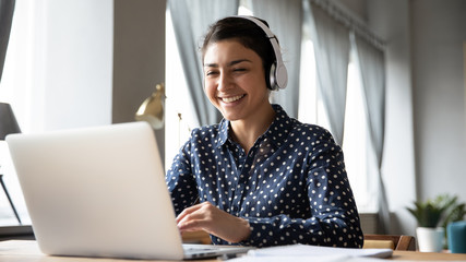 smiling indian girl wearing headphones using laptop, looking at screen, happy young female listening
