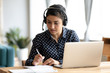 © fizkes - Focused Indian girl wearing headset writing notes in notebook, female student studying language online, listening to lecture, watching trainings, webinar on laptop, sitting at desk