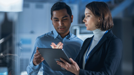 Fototapete - In Technology Research Facility: Female Project Manager Talks With Chief Engineer, they Consult Tablet Computer. Team of Industrial Engineers, Developers Work on Engine Design Using Computers