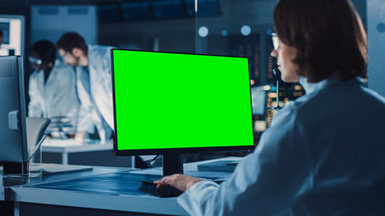 Over the Shoulder Shot: Female IT Scientist Uses Green Mock-up Screen Computer. In the Background Technology Research And Development Laboratory with Scientists, Engineers Working on High Tech Design