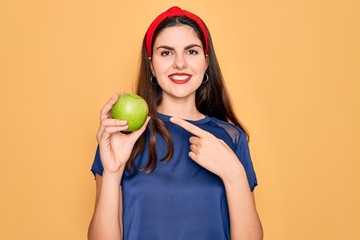 Sticker - Young beautiful girl eating fresh organic healthy green apple over yellow background very happy pointing with hand and finger