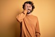 © Krakenimages.com - Young handsome man with beard wearing casual sweater standing over yellow background covering one eye with hand, confident smile on face and surprise emotion.