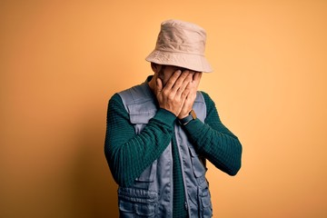 Canvas Print - Handsome tourist man with beard on vacation wearing explorer hat over yellow background with sad expression covering face with hands while crying. Depression concept.