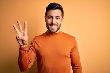 Young handsome man with beard wearing casual sweater standing over yellow background showing and pointing up with fingers number three while smiling confident and happy.