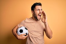 Handsome Player Man With Beard Playing Soccer Holding Footballl Ball Over Yellow Background Shouting And Screaming Loud To Side With Hand On Mouth. Communication Concept.