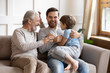 © fizkes - Happy three generations of men sit relax on couch in living room having fun together, overjoyed small preschooler boy play with young dad and elderly grandfather, enjoy family weekend at home