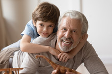 Portrait Of Happy Little Boy Hug Cuddle With Overjoyed Mature Grandfather Playing At Home Together, Smiling Small Preschooler Grandchild Posing Look At Camera Embracing Smiling Senior Grandparent