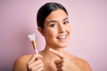 Poster - Young beautiful girl applying cosmetic using spatula over pink background very happy pointing with hand and finger