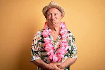 Canvas Print - Grey haired senior man wearing summer hat and hawaiian lei over yellow background puffing cheeks with funny face. Mouth inflated with air, crazy expression.