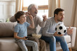© fizkes - Three generations of men sit on couch in living room watch football match game eating popcorn together, little preschooler boy with dad and grandfather cheer team have fun enjoy TV game relax at home