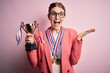 Young beautiful redhead woman holding trophy wearing medals over pink background very happy and excited, winner expression celebrating victory screaming with big smile and raised hands