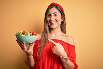 Young beautiful brunette woman eating healthy salad over isolated yellow background very happy pointing with hand and finger