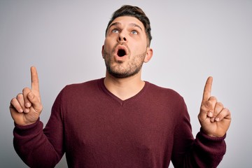 Young man with blue eyes wearing casual sweater standing over isolated background amazed and surprised looking up and pointing with fingers and raised arms.